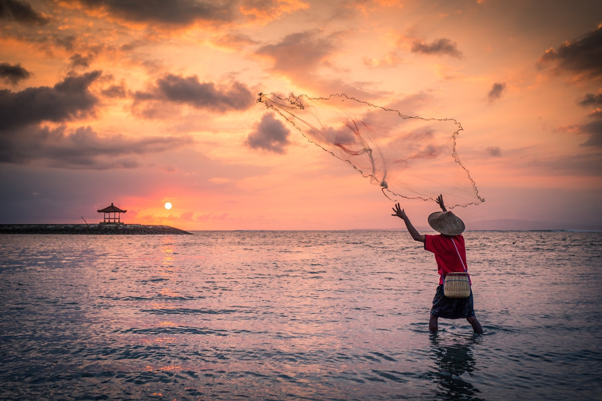 Person casting a net in the ocean at sunset with a pavilion in the background.