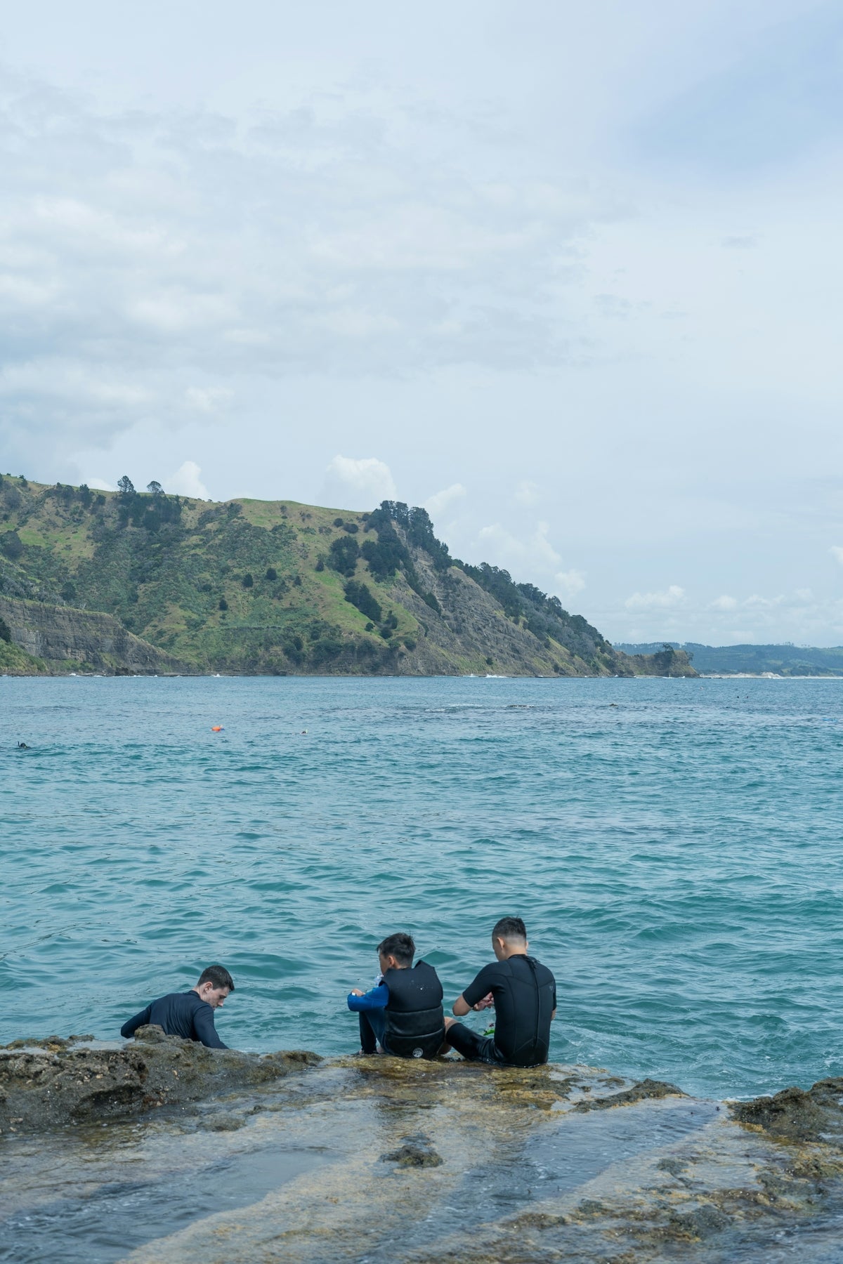 Three people in wetsuits fishing near water with rocky shore and hills in the background.