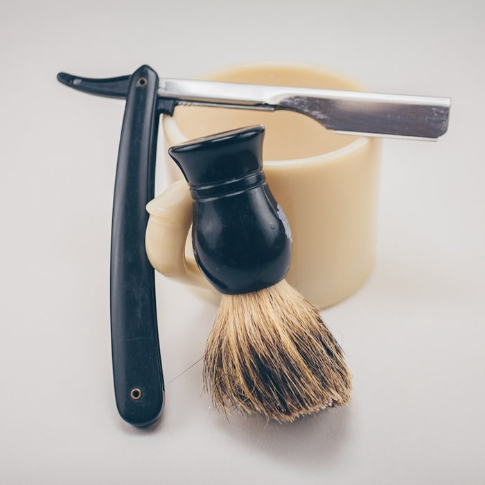 black straight razor beside beige ceramic mug and shaving cream brush