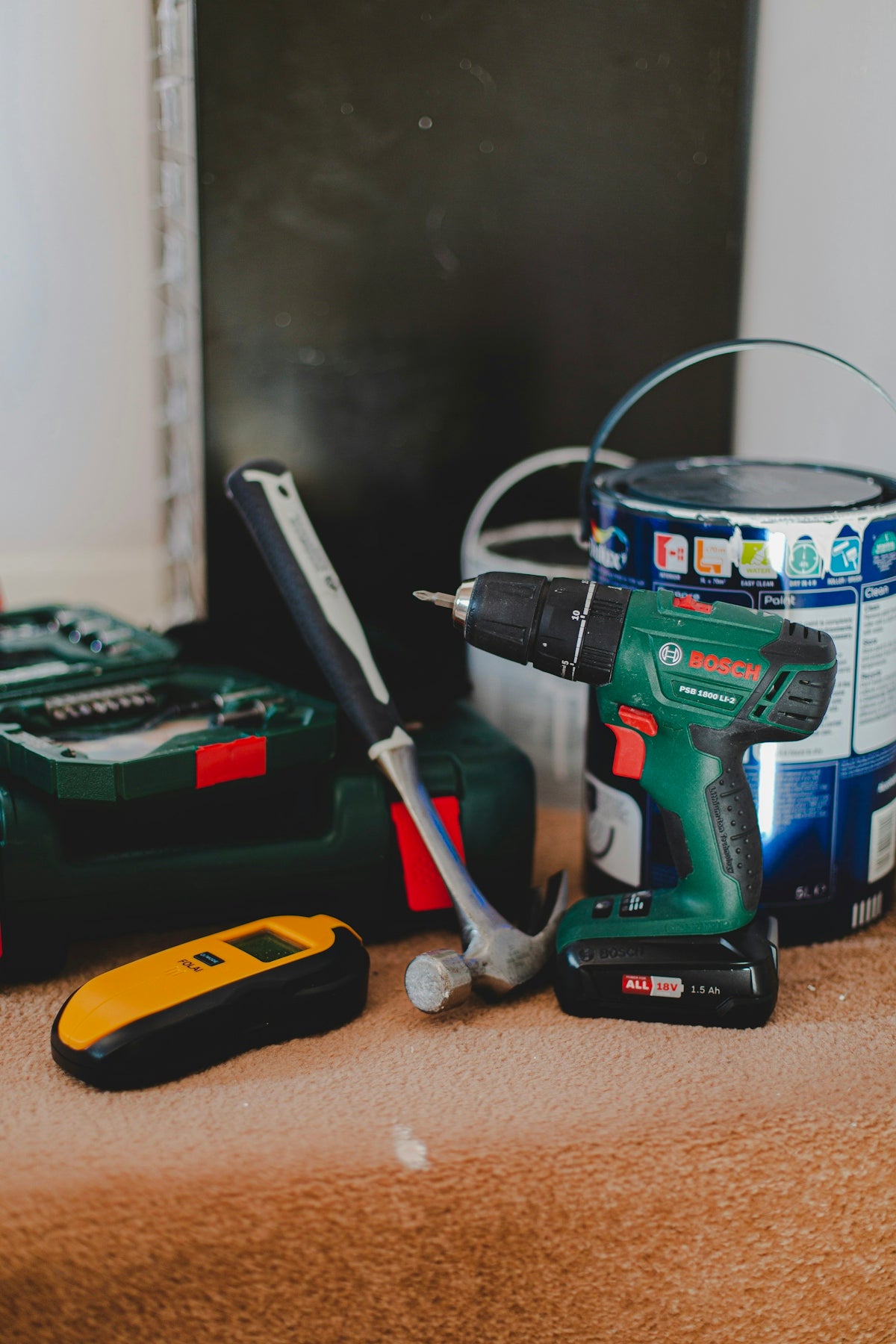 A cordless drill, a hammer, and a yellow stud finder on a carpeted floor