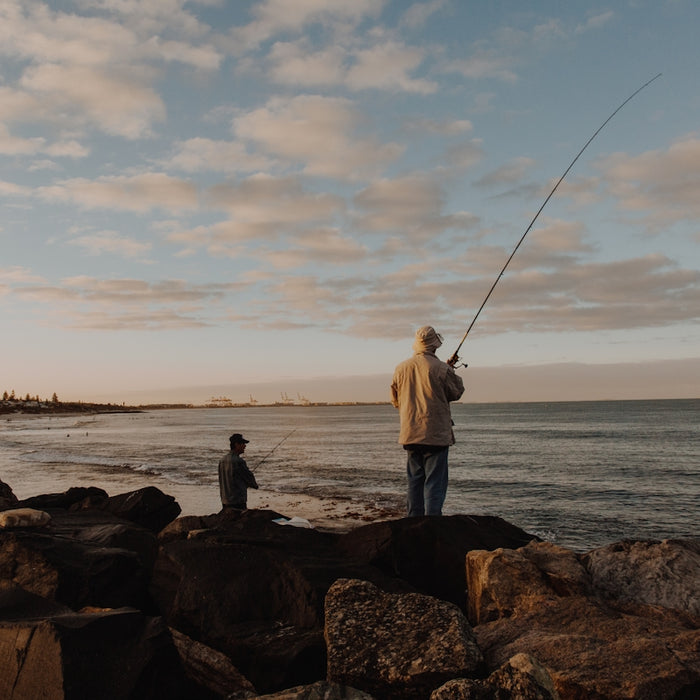 man in white shirt and black pants holding fishing rod standing on rock near sea during