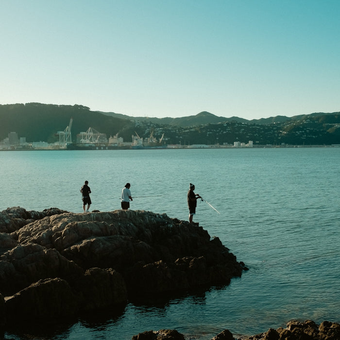 Three people fishing on rocks by a calm bay.