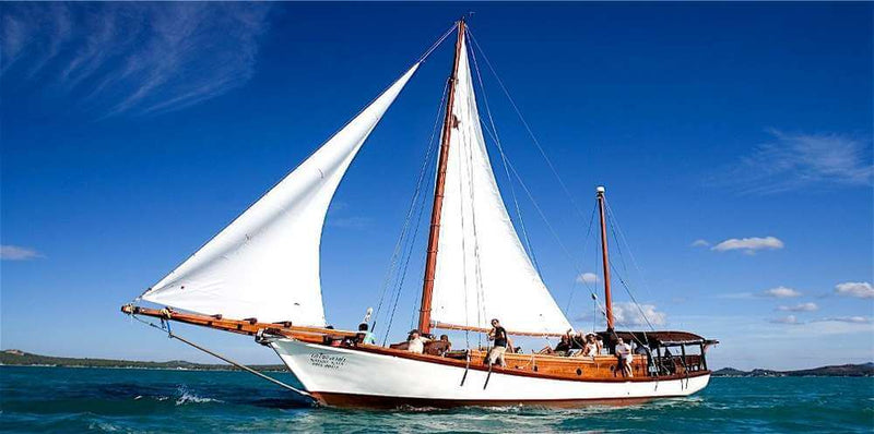 Sailboat on clear waters with a blue sky, showcasing a classic wooden design and multiple people on board.